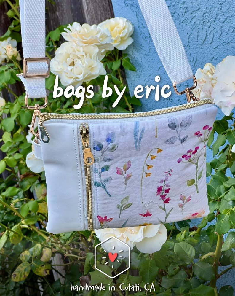 A handmade floral crossbody bag with a white adjustable strap and gold-colored zippers, hanging among white flowers.